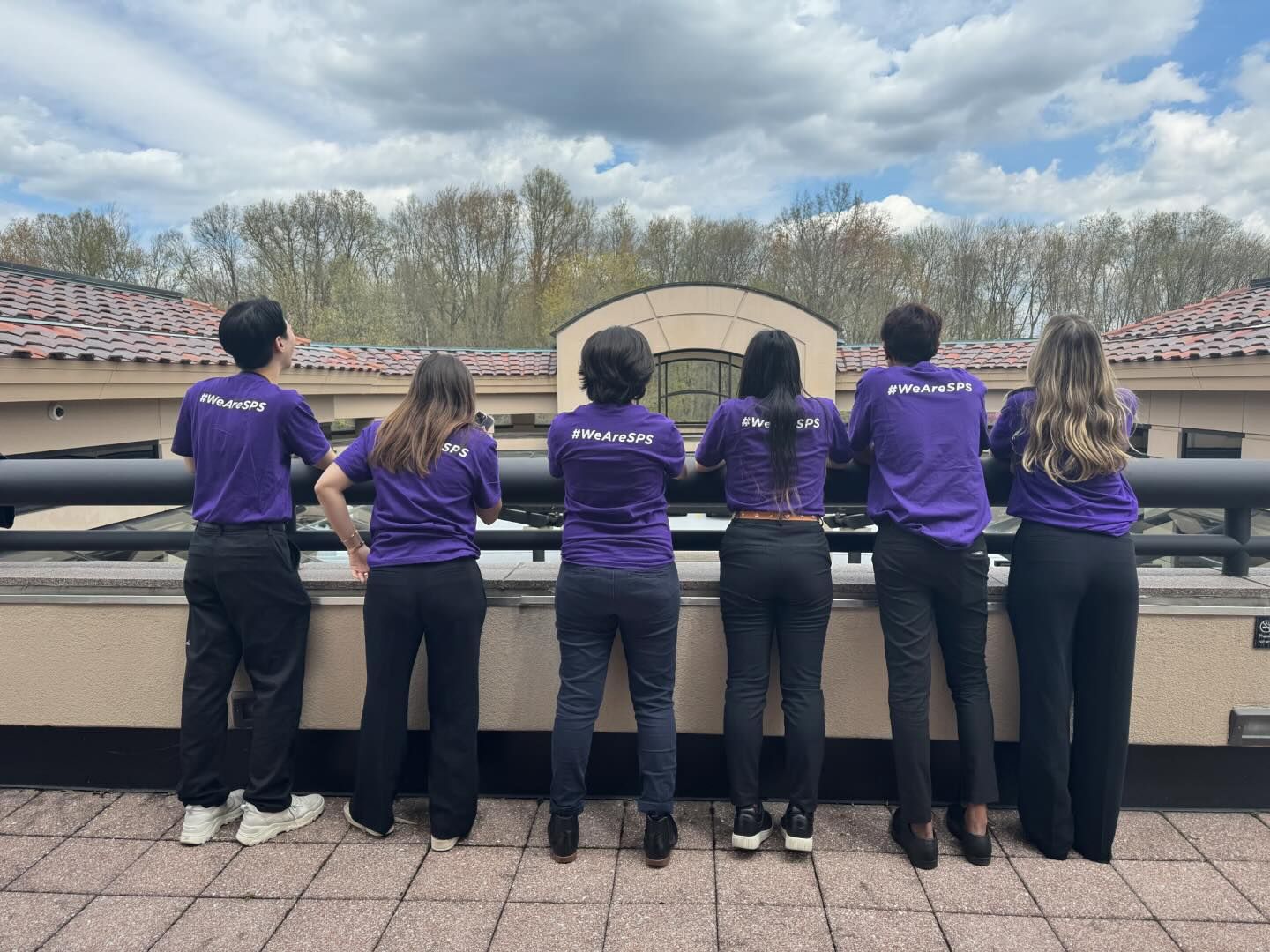 group of college aged students facing away from the camera showing matching shirt backs while standing on balcony